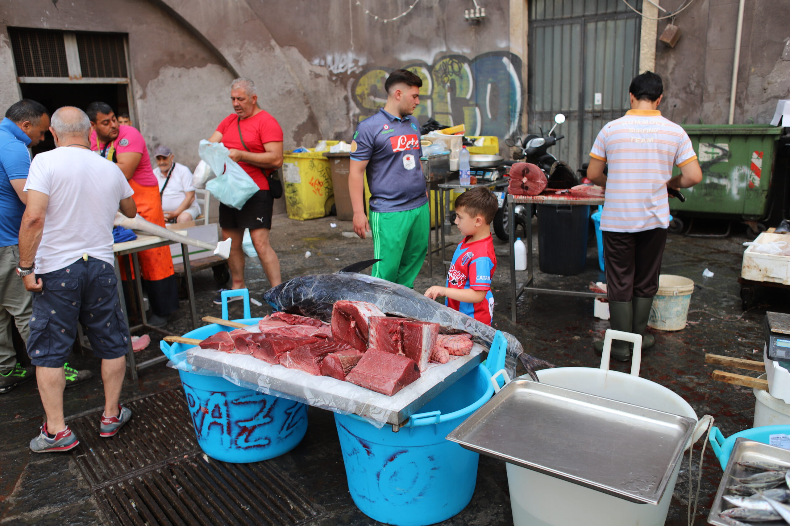 A child stands next to a large container filled with neatly cut fish pieces, their vibrant red color contrasting with the blue of the container. The boy's bright jersey adds a touch of innocence and curiosity to the scene. Other vendors, seen in the background, continue their work, surrounded by various bins and crates. The mix of generations highlights the market's role in the community, bridging past and future through daily interactions and traditions.