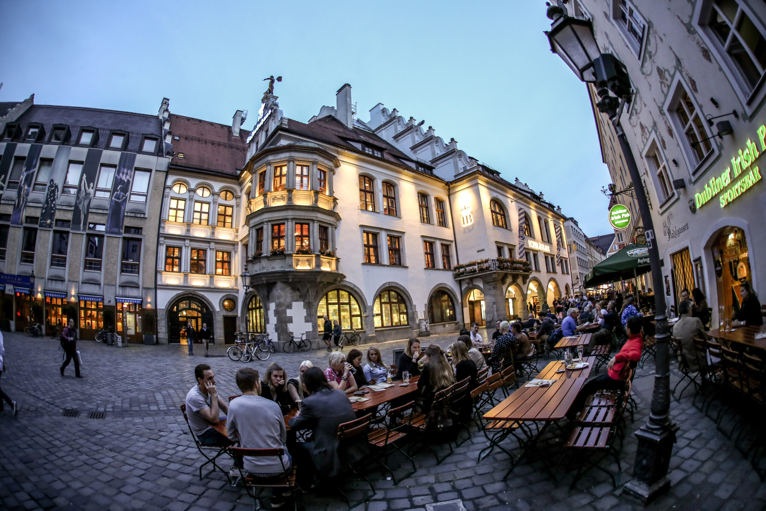 The Platzl in Munich at dusk, the lit facade of the Hofbrauhaus above long beer garden tables full of people