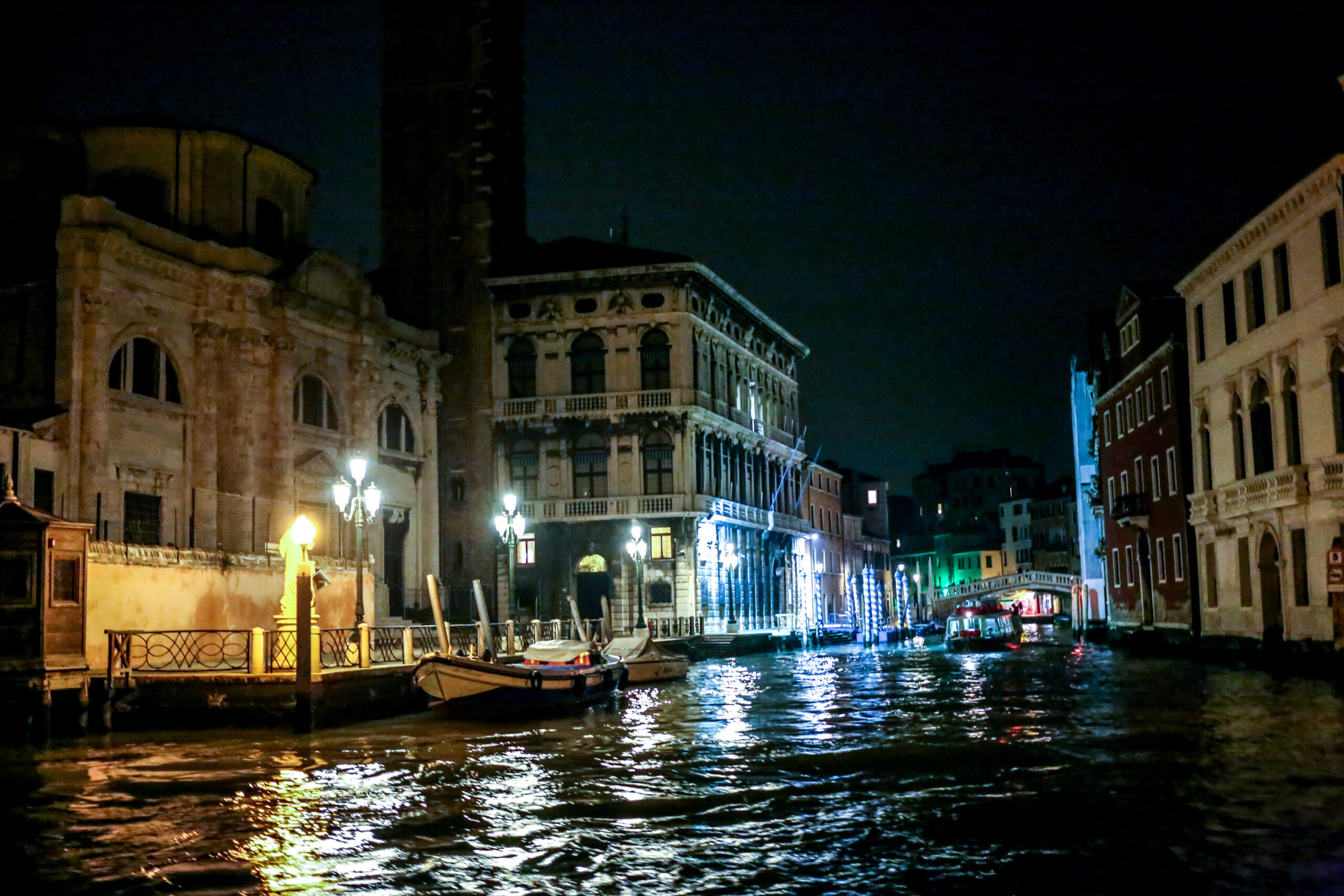 A Venetian canal at night seen from the water, lamps and mooring poles reflected in the ripples beside a lit palazzo