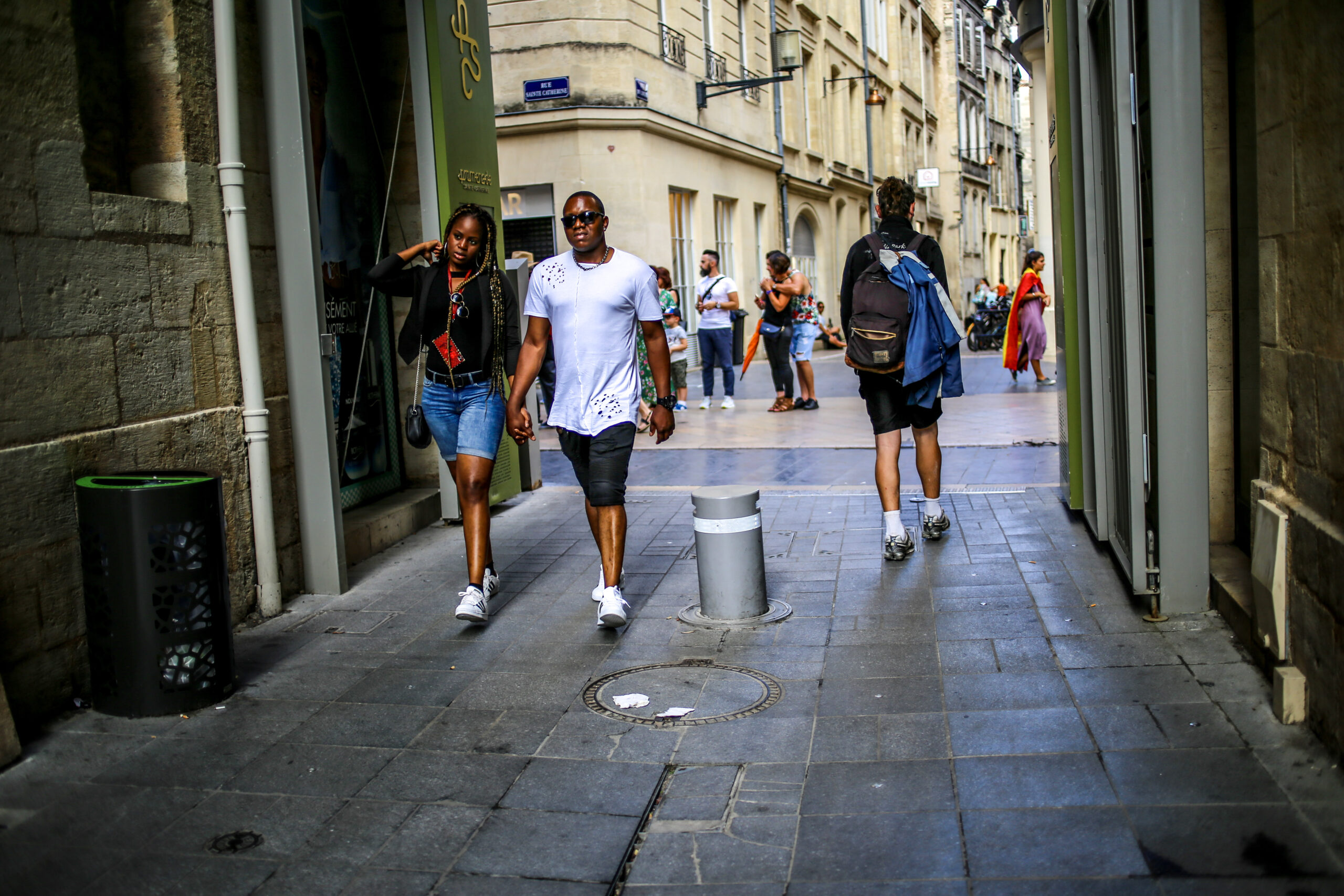 A shaded side lane off Rue Sainte-Catherine in Bordeaux looking out onto the sunlit shopping street, with a couple walking hand in hand toward the camera