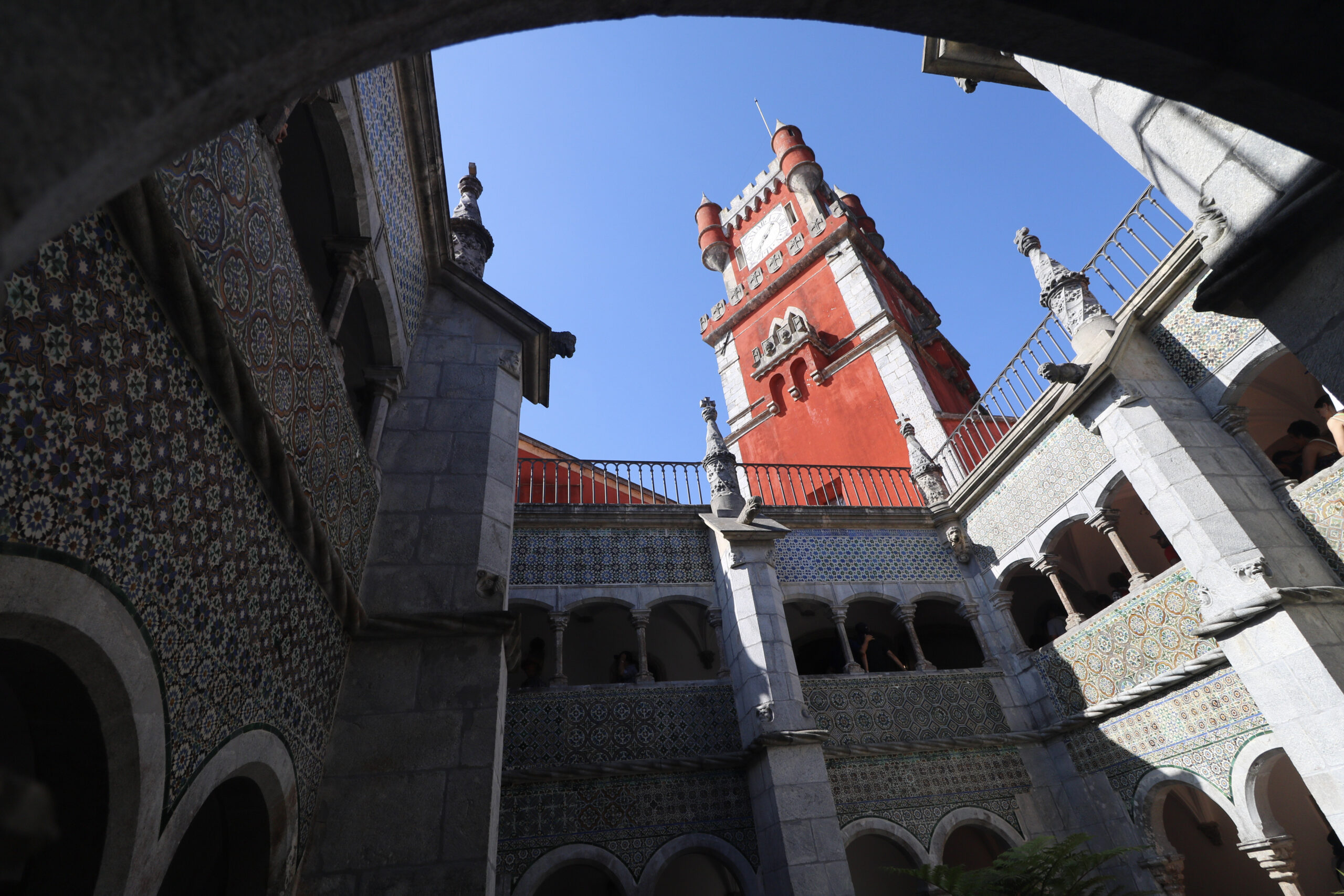 The tiled cloister of Pena Palace in Sintra, looking up past azulejo walls to the red clock tower against a deep blue sky