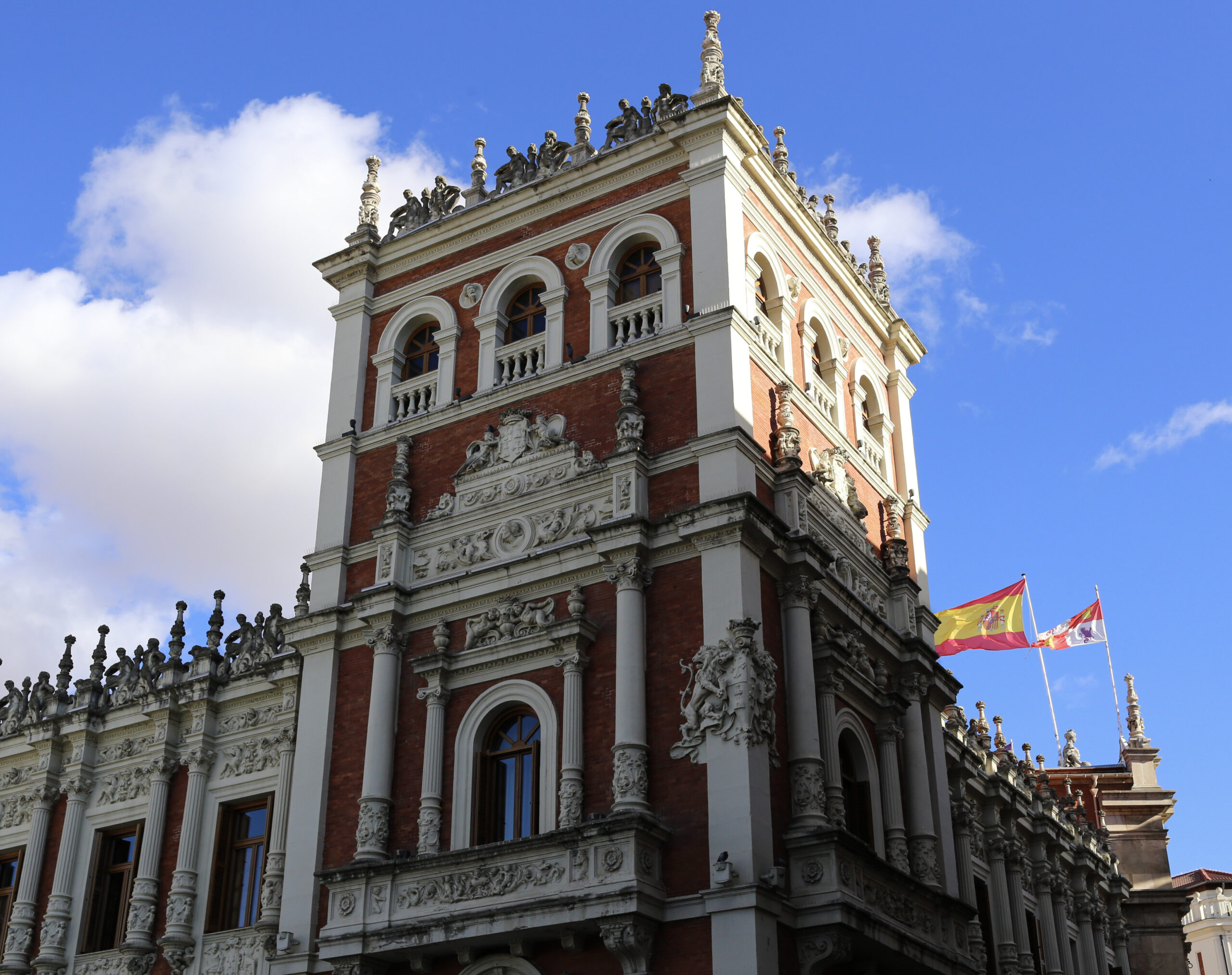 The upper storeys of an ornate brick and white stone civic palace in Castile and Leon, statues along the parapet, Spanish and Castilian flags flying