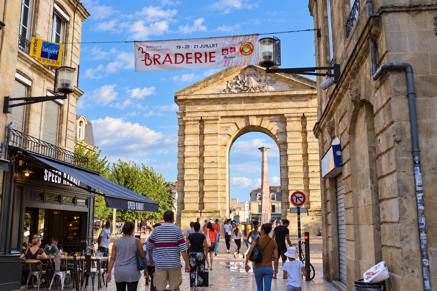 Pedestrians and cafe tables on a Bordeaux shopping street, with a summer sale banner above the sunlit Porte Dijeaux.