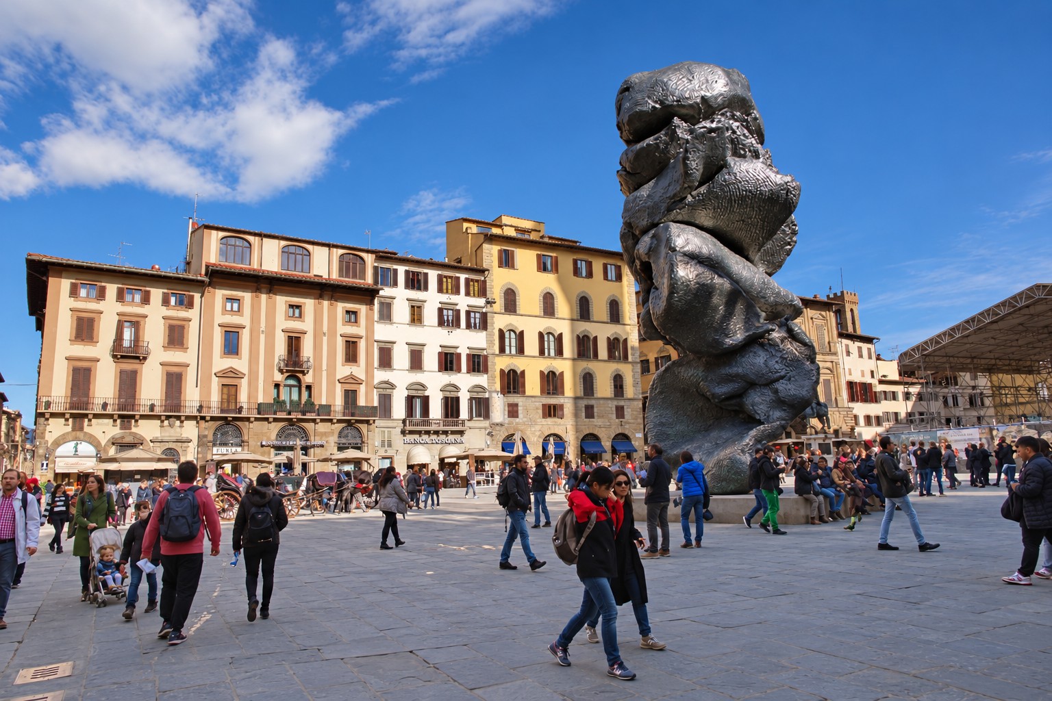 Visitors cross Piazza della Signoria beneath Urs Fischer's towering Big Clay sculpture, framed by warm historic facades and a blue sky.