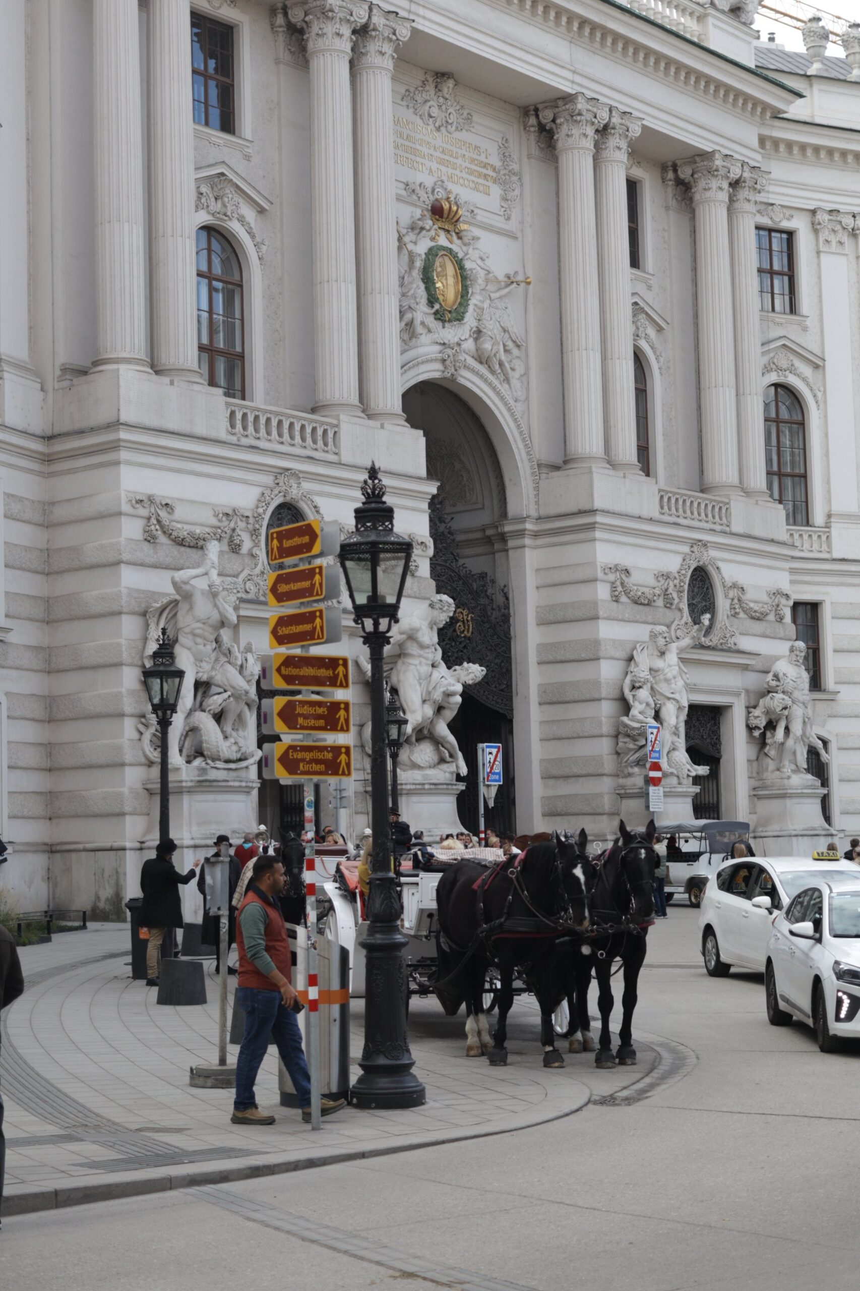 The Michaelertor gate of the Hofburg on Michaelerplatz in Vienna, with the Franz Joseph inscription and crown above the arch, marble Hercules statue groups either side and a Fiaker carriage waiting