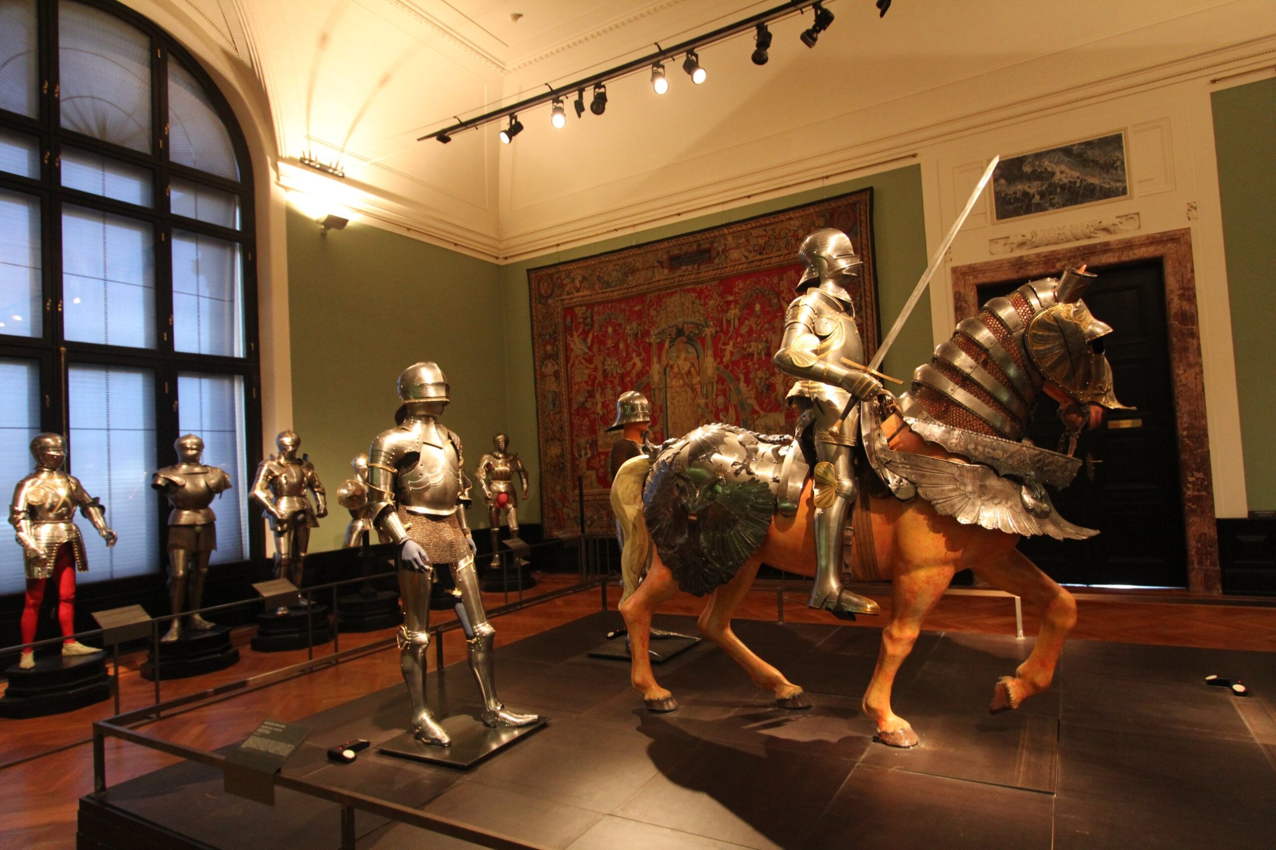 A knight with a raised sword on a horse in full plate horse armour, with standing armours by a tall window and a red tapestry behind, in the Imperial Armoury in Vienna