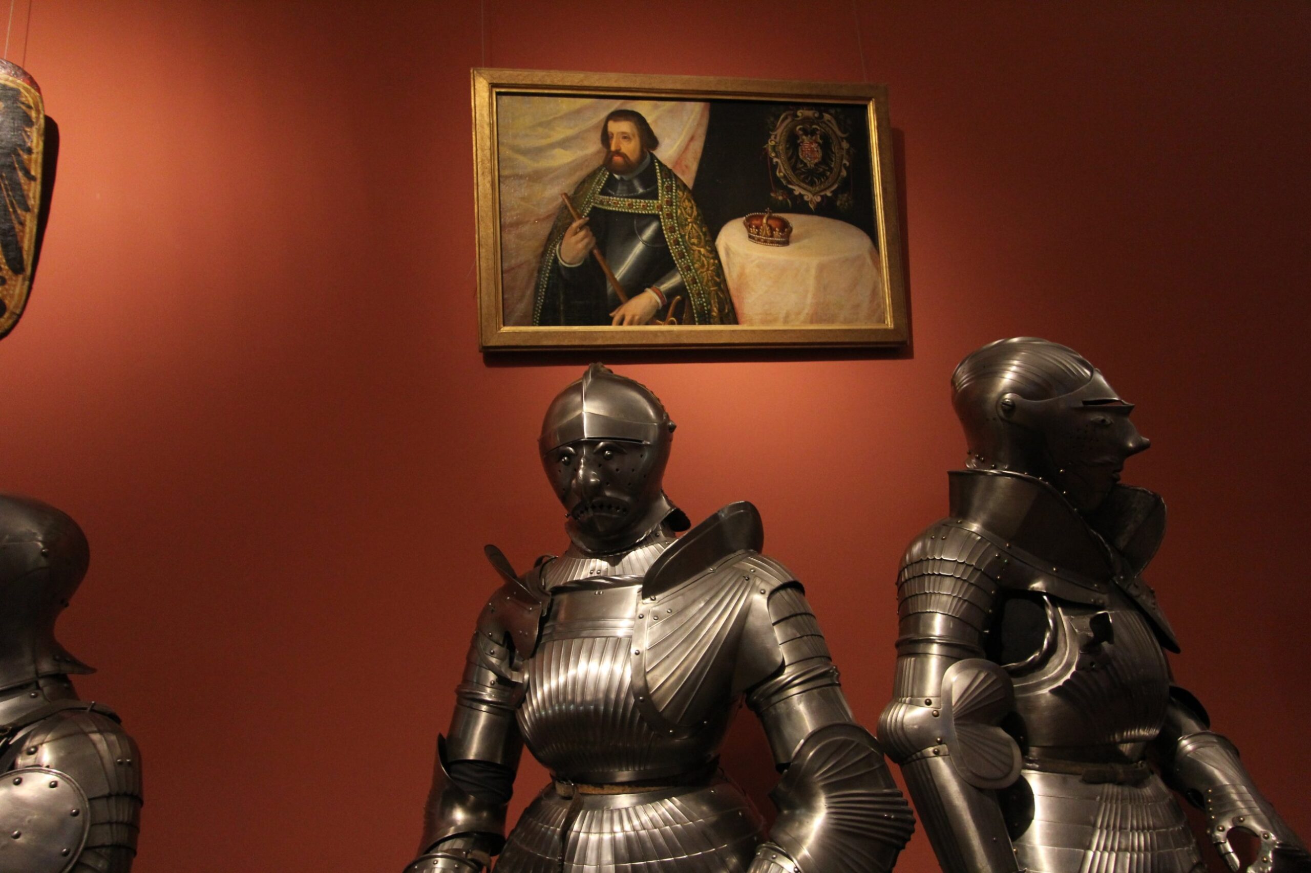 Fluted steel armours, one with a visor shaped as a grotesque human face, below a portrait of an armoured ruler in the Imperial Armoury in Vienna