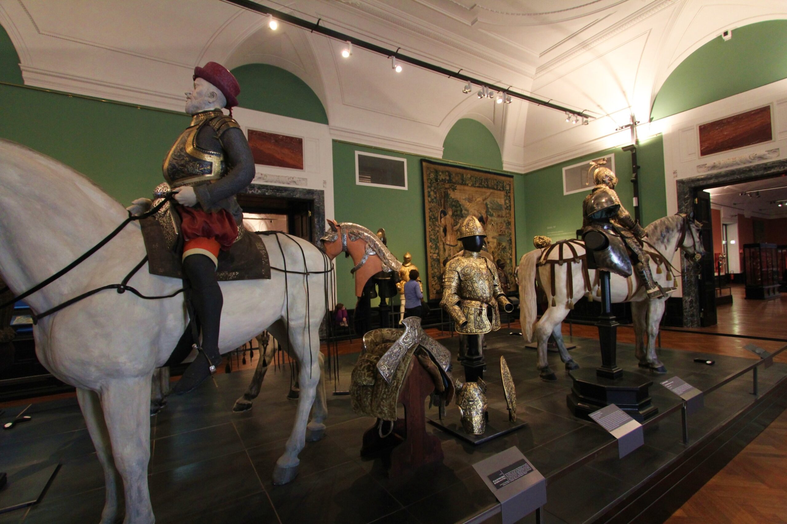 Mounted figures and gilded parade armours on white horses in a green-walled room of the Imperial Armoury in Vienna