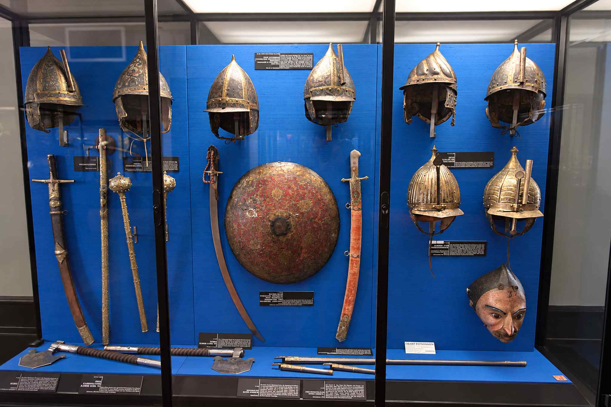 A blue display case of pointed Ottoman-style helmets, a red and gold round shield, maces, sabres and a painted face mask in the Imperial Armoury in Vienna