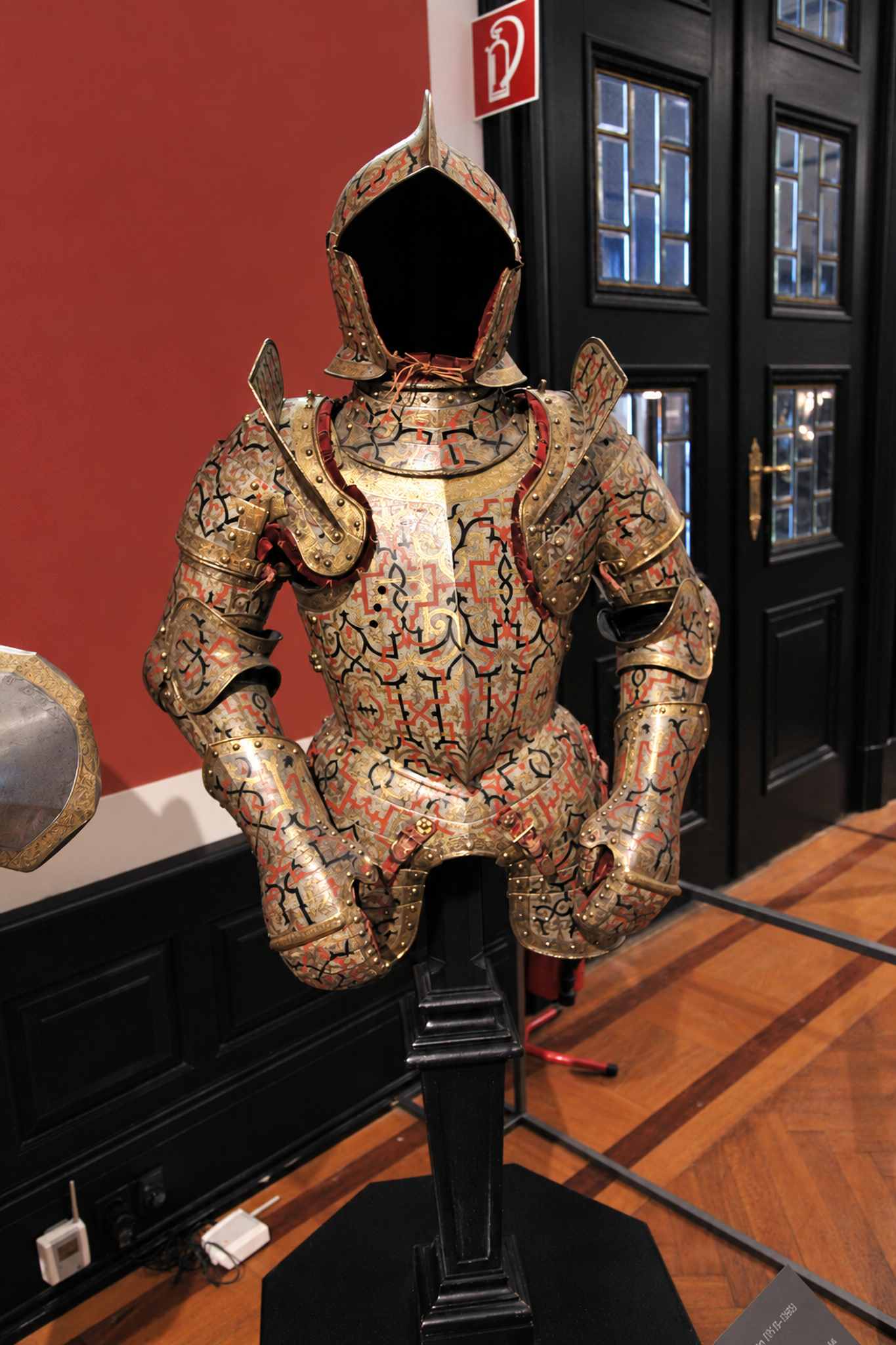 A half armour and open helmet covered in red, black and gold strapwork on a stand in the Imperial Armoury in the Neue Burg in Vienna