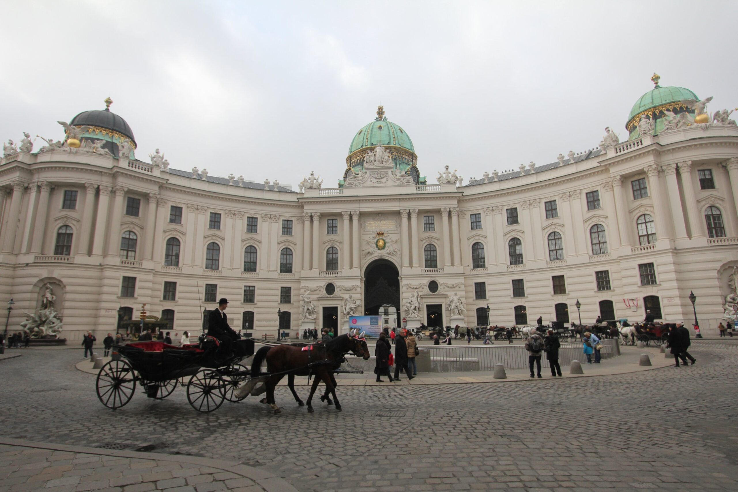 A Fiaker with a coachman in a bowler hat driving past the curved Michaelertrakt of the Hofburg and its green dome on Michaelerplatz in Vienna, with more carriages waiting at the stand by the Michaelertor