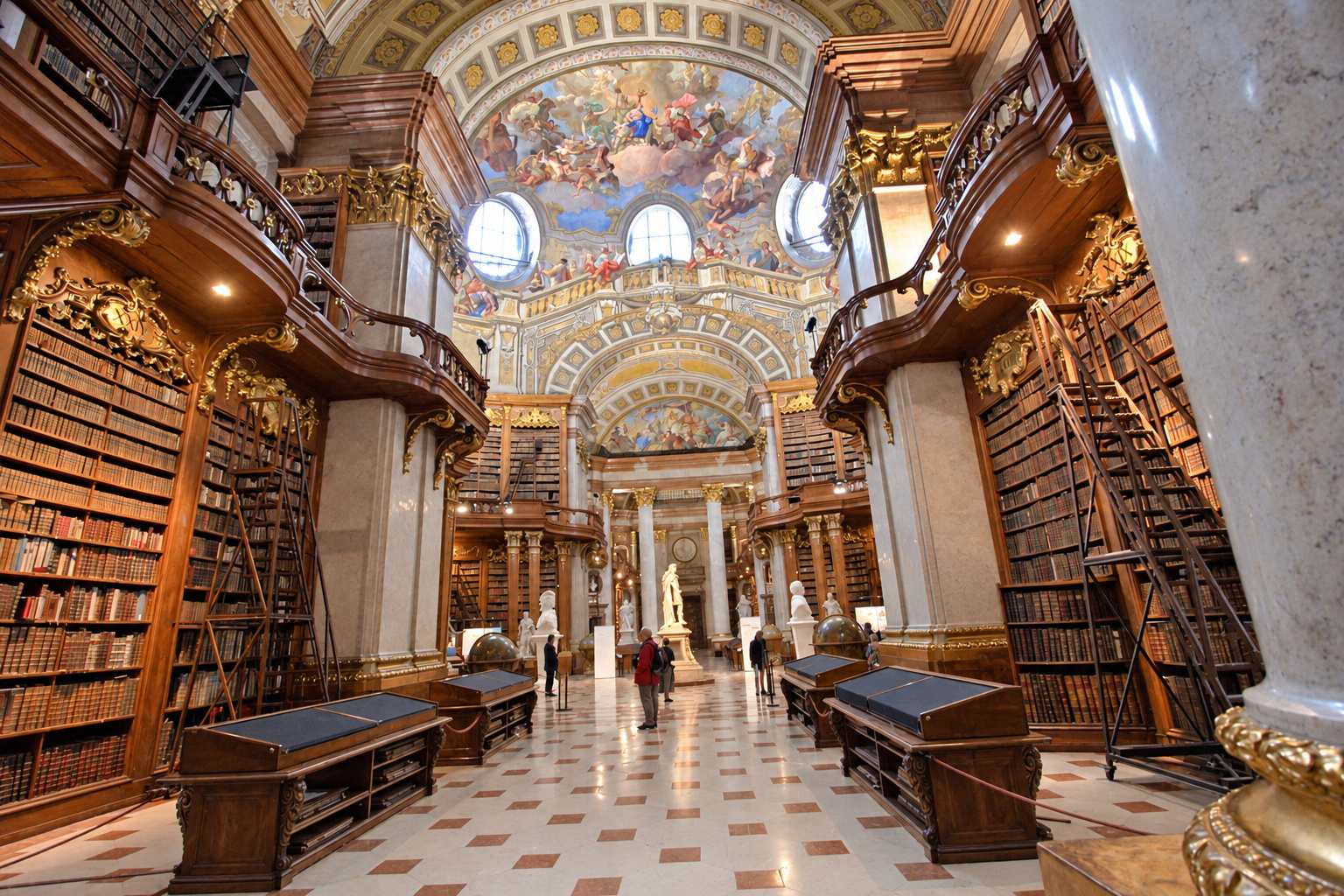 Tall wooden bookcases and curving galleries frame a view toward frescoes and a central statue in Vienna's State Hall.